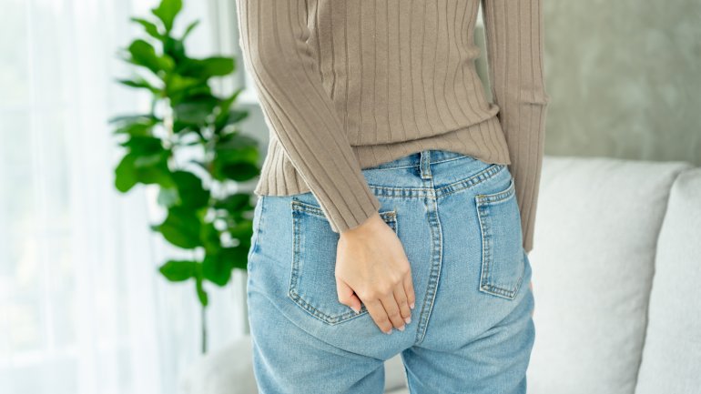 Eine Frau in Jeans und Pullover hält ihre linken Hand an den Po.